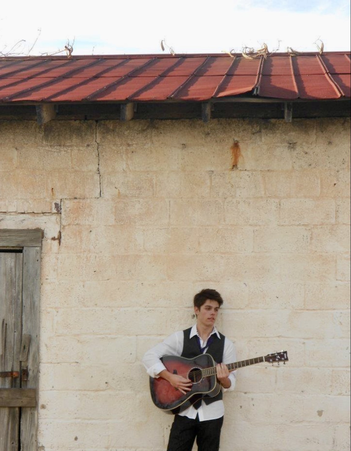 Julius Cole Rassin with acoustic guitar, standing against a whitewashed stone wall under a red metal roof