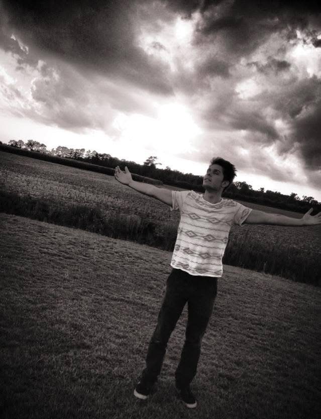 Julius Cole Rassin standing in a field with arms outstretched beneath a dramatic cloudy sky, black and white