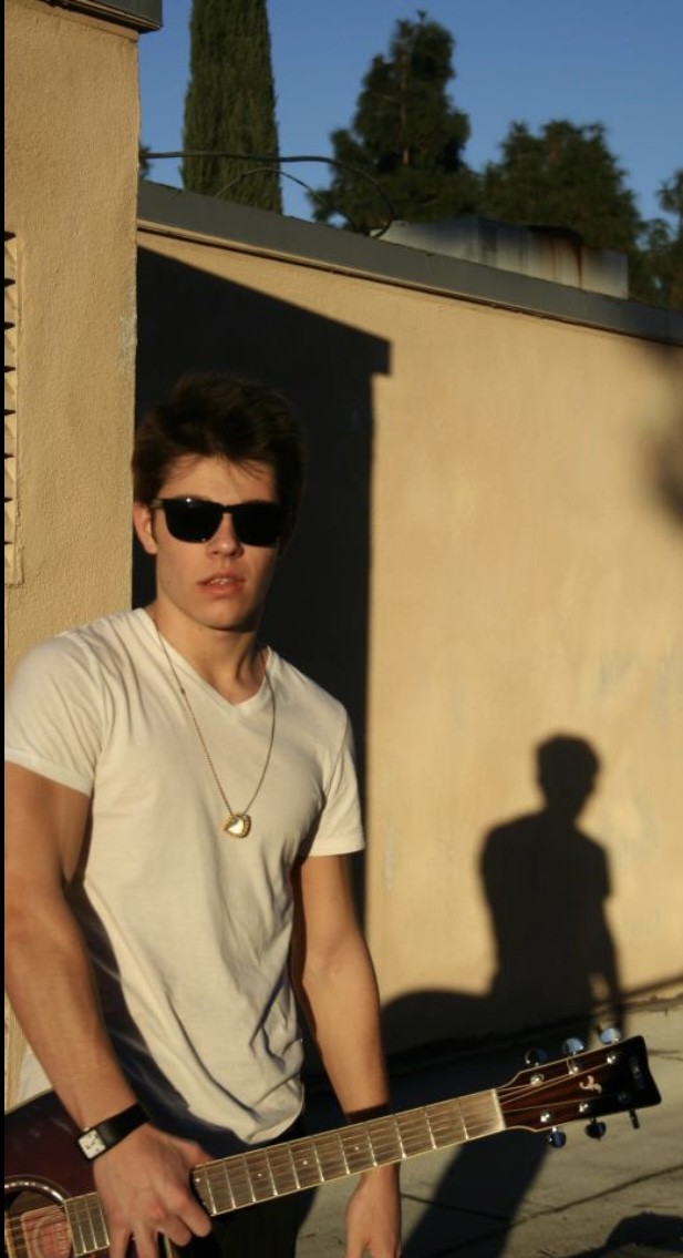 Julius Cole Rassin in a white t-shirt and sunglasses holding an acoustic guitar in golden afternoon light