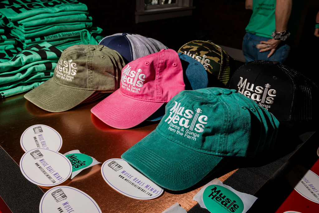 Music Heals merchandise table at the 2025 fundraiser: caps in multiple colors with the 'Stories & Songs from the Farm' wordmark